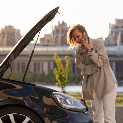 Woman driver calling for help assistance looking at broken car with opened hood, talking on mobile
